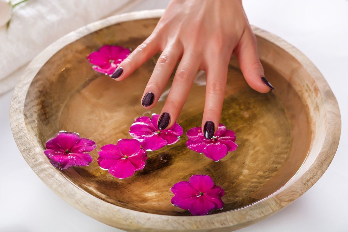 Female hand with purple varnish on nails touching flower in wooden bowl with water. Manicure and Spa beauty treatment concept. Selective focus, close up